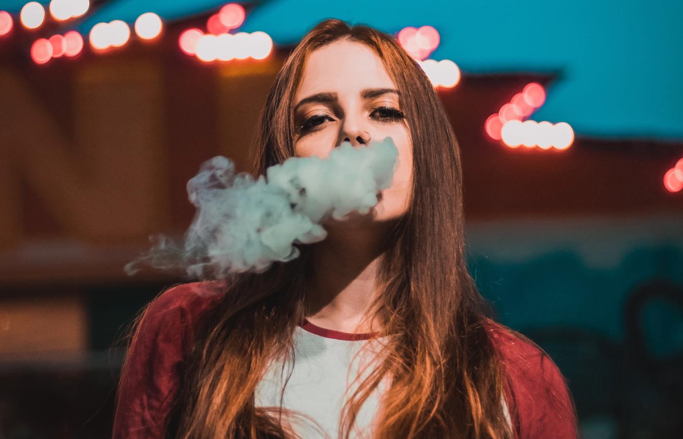 Młodzi ludzie na imprezie, jeden z nich podaje drugiemu kolorowy, jednorazowy e-papieros, symbolizujący pierwszy, niewinny kontakt.