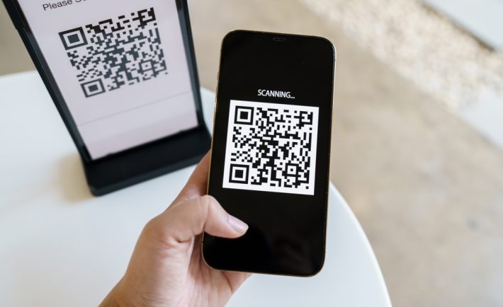 Ręka trzymająca smartfon skanujący kod QR na opakowaniu produktu Fumot Vape, z ekranem telefonu pokazującym zielone potwierdzenie autentyczności.