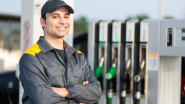 Pracownik stacji benzynowej, w tle dystrybutory paliwa i ekspres do kawy, z dezorientacją lub brakiem wiedzy patrzy na e-papieros, podczas gdy klient czeka na obsługę.