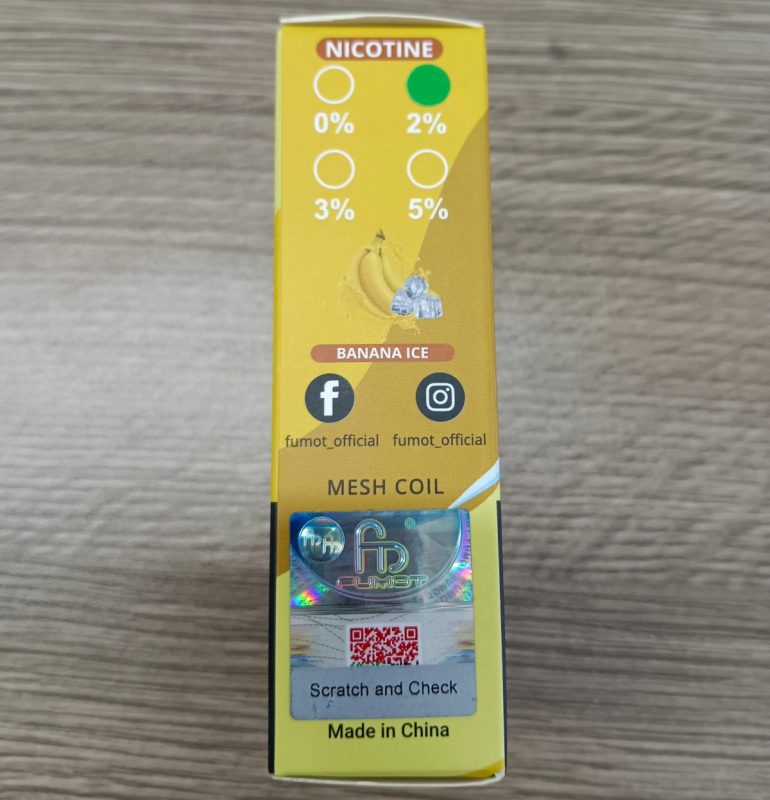 Zbliżenie na oryginalne opakowanie e-papierosa Fumot Tornado 15000, pokazujące błyszczące hologramy, wytłoczenia i wysokiej jakości teksturę papieru pod światło.