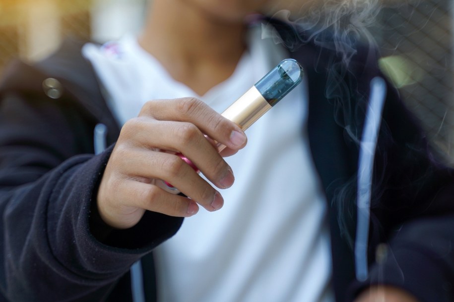 Nastolatek wapujący e-papierosa, z sylwetką tradycyjnego papierosa niewyraźnie widoczną w tle, symbolizującą